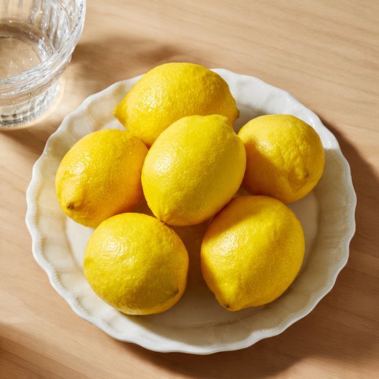 The image shows a square baking pan lined with white parchment paper, filled with a smooth, shiny yellow batter layer that looks thick and spread evenly across the pan. The background surface is a white marbled texture, and there is soft natural light coming from the left side, casting subtle shadows on the batter and pan edges. Photo taken with an iphone --ar 4:5 --v 7