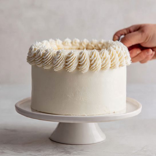 A round cake covered in smooth white cream frosting sits on a white cake stand, with a white marbled texture in the background. On top, there is a decorative border made of evenly spaced, curved layers of piped white frosting with ridged texture, formed by a metal piping tip held by a woman's hand that is visible in the top right corner. The cake shows a simple, clean look with a soft creamy texture that covers the whole surface evenly. Photo taken with an iphone --ar 4:5 --v 7