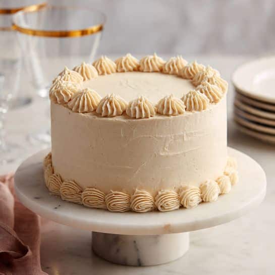 A round two-layer cake is covered smoothly in light cream-colored frosting with a soft texture. The top and bottom edges are decorated with evenly spaced small swirls of the same cream-colored frosting. The cake sits on a white marble cake stand with a thick base, set on a white marbled surface. In the background, there are clear glasses with thin gold rims and a stack of white plates. The overall look is clean and simple with soft lighting. photo taken with an iphone --ar 4:5 --v 7