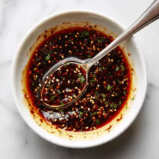 Two long oval pieces of dough lie on a wooden board with a light dusting of flour. Both dough pieces have a thick layer of greenish-brown paste spread unevenly on top, filled with many small seeds, giving a textured look. A shiny metal spoon covered with this seed-filled paste rests on the left piece of dough. The dough is soft and slightly puffy around the edges, creating a clear border around the paste. The background is a white marbled texture. photo taken with an iphone --ar 4:5 --v 7