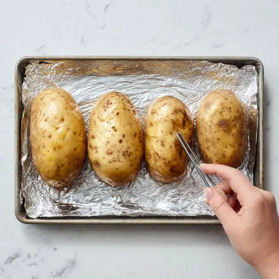The image shows four large light brown potatoes with rough skin lying on a baking tray lined with shiny silver aluminum foil. The potatoes have small, scattered dark spots and are placed in a slightly uneven row. A woman's hand is holding a metal tool, removing some skin or soft spots from the closest potato. The baking tray rests on a white marbled surface, giving a clean and bright background. Photo taken with an iphone --ar 4:5 --v 7