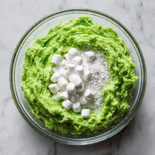 A clear glass bowl filled with three layers: the bottom layer is a pale green creamy mixture, the middle layer is slightly visible and appears smooth, topped with a thick layer of small white mini marshmallows that cover the green base fully. A black spatula is partially inserted on the right side of the bowl, resting inside the mixture. The bowl is placed on a white marbled surface with parts of two other white bowls visible at the top edges. photo taken with an iphone --ar 4:5 --v 7