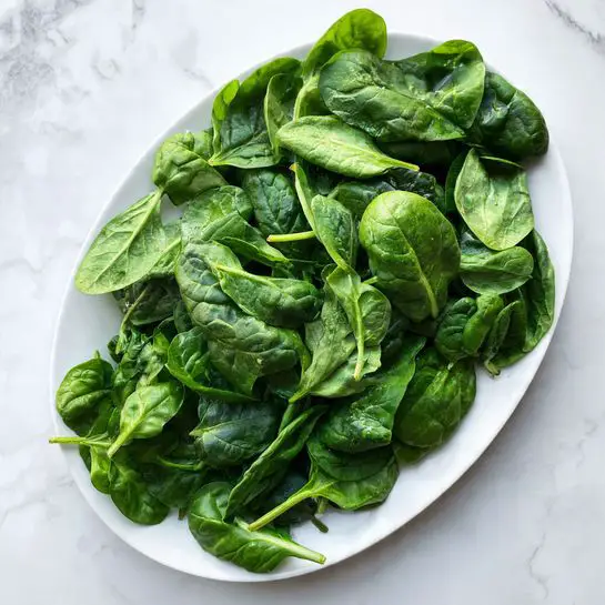 An oval white plate filled with fresh spinach leaves piled high, showing many different shades of green and smooth textured leaves with visible veins and stems. The plate sits on a white marbled surface. photo taken with an iphone --ar 4:5 --v 7
