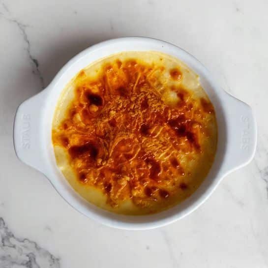 A round white ceramic dish with two handles shows a single layer of crème brûlée on a white marbled surface. The top layer is a caramelized golden-brown sugar crust with some darker caramel spots, giving it a slightly rough texture. The custard beneath is smooth, creamy, and pale yellow, visible through the subtle gloss of the caramelized sugar on top. The dish is centered in the frame, and the handles have the word