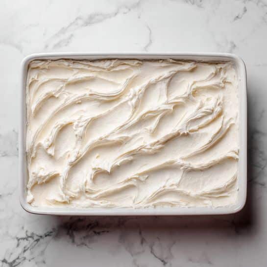 The image shows a white rectangular dish filled with a dessert that has two visible layers. The bottom layer consists of small pieces of light beige cake or bread, soaked slightly. The top layer is a thick, shiny, bright red cherry topping with whole cherries, covering the entire surface unevenly, creating a glossy and juicy texture. The dish sits on a white marbled surface, with natural light highlighting the rich red color and the texture of the cherries. photo taken with an iphone --ar 4:5 --v 7