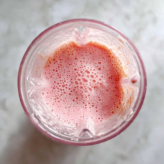 A top-down view inside a clear blender shows a single layer of light pink smoothie with small bubbles on the surface, with a few small bits clinging to the blender walls. The blender is placed on a white marbled surface. photo taken with an iphone --ar 4:5 --v 7
