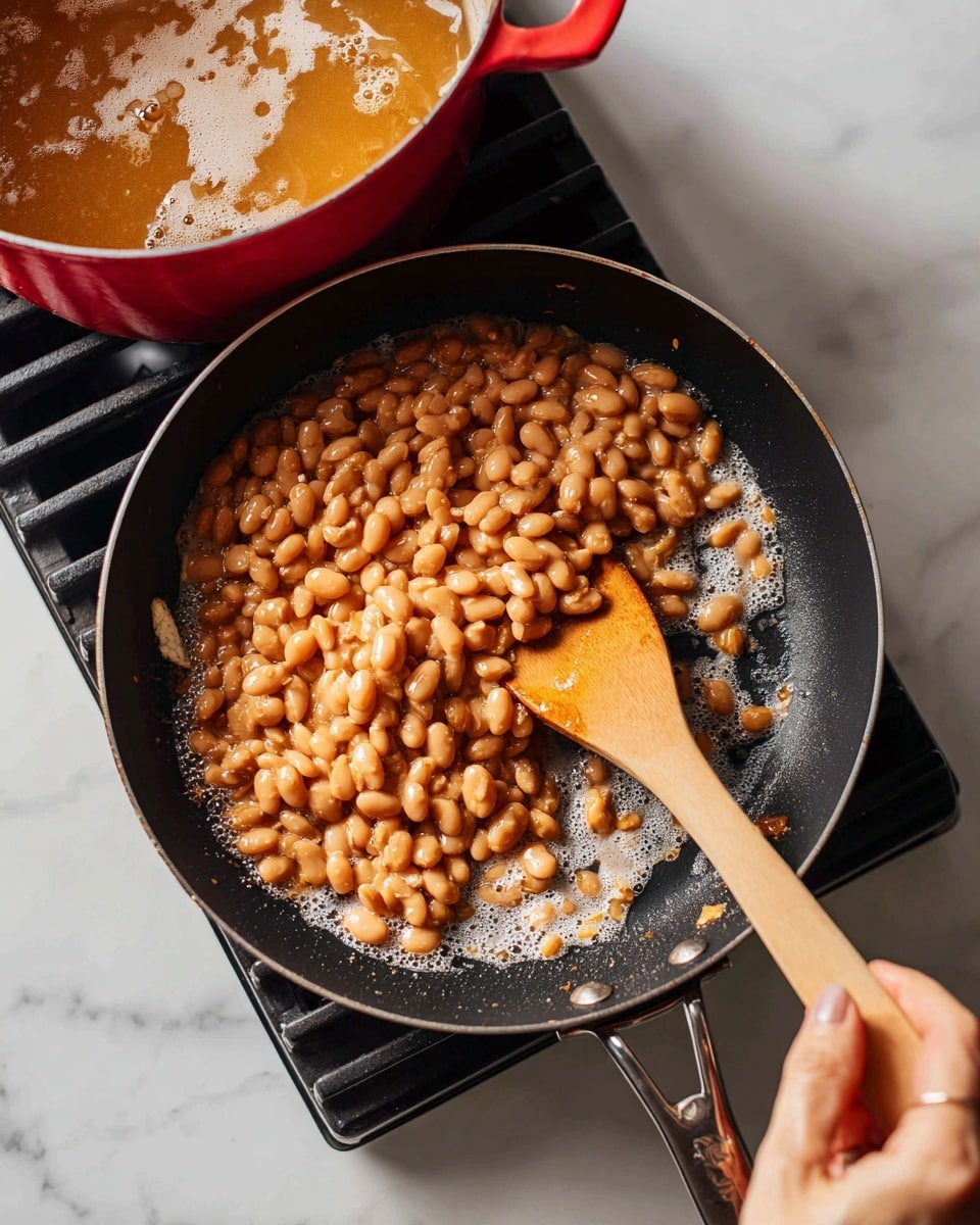A close-up view of a black frying pan on a stovetop filled with cooked light brown beans, some broken open, creating a rough texture. A wooden spatula held by a woman's hand is stirring the beans from the right side. Behind the frying pan is a red pot with more beans soaking in a cloudy liquid. The stovetop is black with metal grates, and the surface below has a white marbled texture. photo taken with an iphone --ar 4:5 --v 7