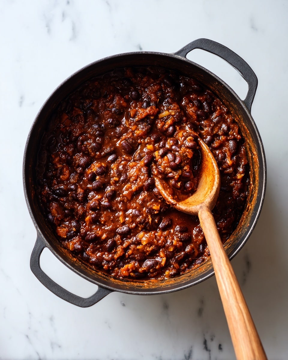 Inside a black pot, there is a thick, chunky mixture that looks like cooked beans with mashed texture. The colors are mostly dark brown and black, with some orange-brown bits showing the cooked ingredients. A wooden spoon rests on the right side, partly dipped into the mix, showing its rough, cooked texture. The pot sits on a white marbled surface. photo taken with an iphone --ar 4:5 --v 7