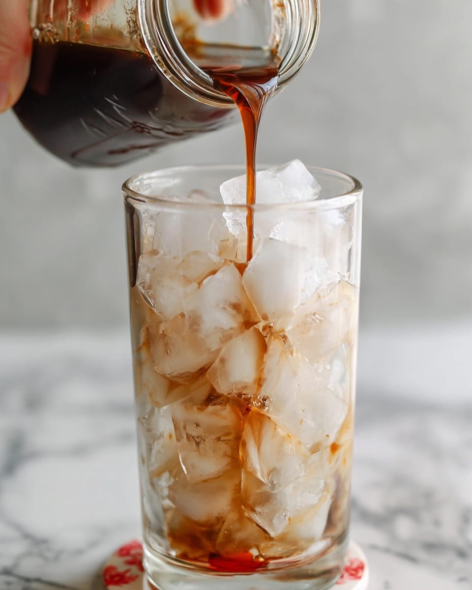 A clear glass filled with three visible layers sits on a white saucer on a white marble surface; the bottom layer is a bright red syrup, above it is a layer of clear ice cubes, and the top layer shows white milk being poured into the glass, swirling with light brown coffee creating a marbled effect. A woman's hand holding the bottle of milk is visible at the top left, pouring the milk. The background is softly blurred with hints of white and red objects. Photo taken with an iphone --ar 4:5 --v 7