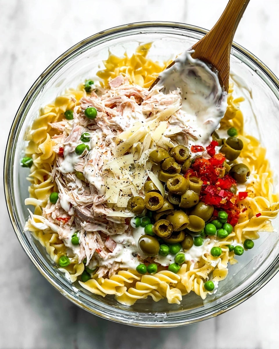A clear glass bowl holds four main layers of food, placed on a white marbled surface: the bottom layer is yellow cooked curly egg noodles; above it is a creamy white sauce spread unevenly; on top of the sauce are bright green peas, light pink shredded cooked meat, sliced green olives, small pieces of red peppers, and a sprinkle of shredded white cheese and black pepper; a wooden spoon with white sauce on its side rests in the bowl from the top right. Photo taken with an iphone --ar 4:5 --v 7