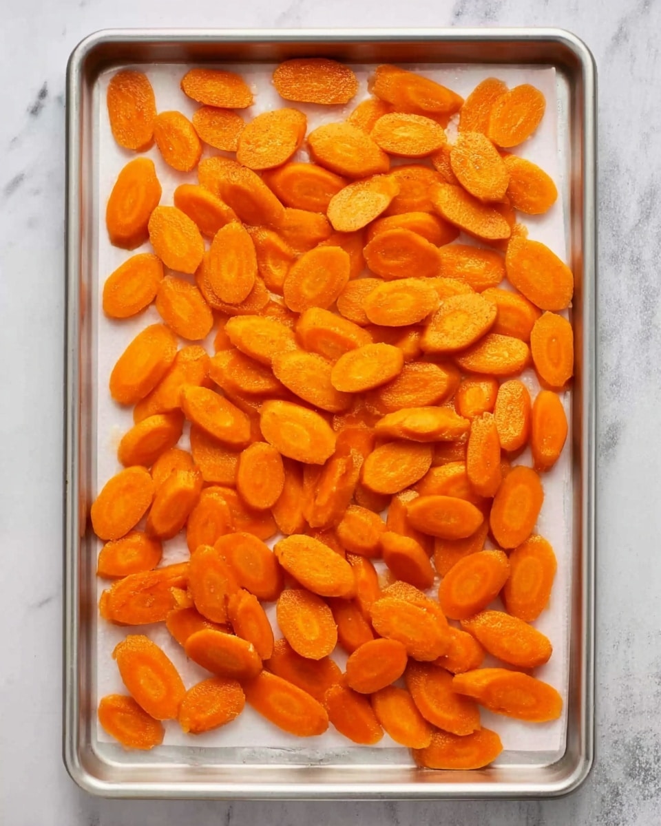 The image shows a silver baking tray lined with white baking paper filled with sliced orange carrots. The carrot pieces are smooth and slightly shiny, arranged in a loose, even layer covering the entire tray. The surface beneath the tray is white with a soft marbled texture, giving a clean and bright look to the scene. Photo taken with an iphone --ar 4:5 --v 7
