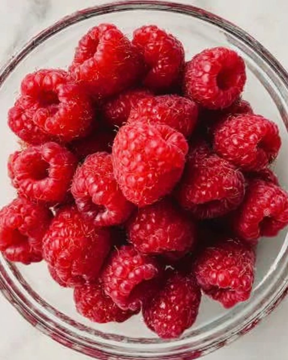 Two metal muffin trays filled with dark brown chocolate batter, each cavity roughly two-thirds full with thick textured batter. In the bottom tray, each batter-filled cup is topped with a bright red raspberry centered on top. A woman's hand is placing a raspberry on one of the batter cups in the bottom tray. Both trays rest on a white marbled surface, with the contrast between the dark chocolate batter and the bright raspberries clearly visible. Photo taken with an iphone --ar 4:5 --v 7