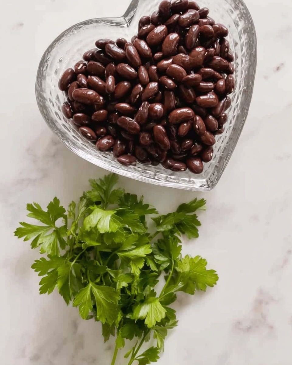 The image shows a clear glass, heart-shaped bowl filled with dark brown beans that have a smooth and shiny texture, sitting on a white marbled surface. Below the bowl, there is a small bunch of fresh green parsley with detailed leaves and a natural texture, lying flat on the same surface. The colors contrast well, with the dark beans and bright green parsley vibrant against the white marble background. photo taken with an iphone --ar 4:5 --v 7