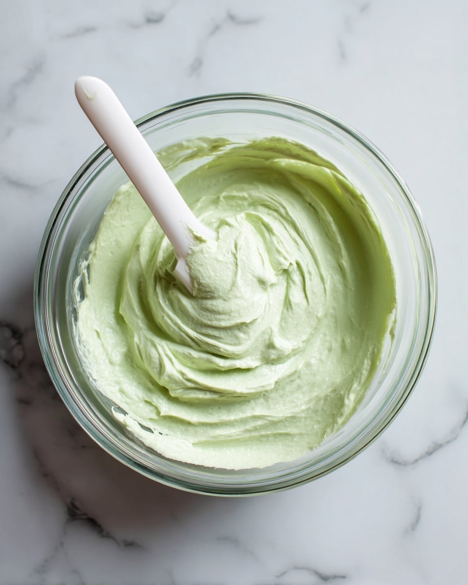 A clear glass bowl with a smooth, thick light green creamy mixture inside. A white spatula is partially dipped into the mixture, with the spatula head coated in the green cream. The bowl sits on a white marbled surface, and the inside texture looks smooth with some gentle swirls made by mixing. Photo taken with an iphone --ar 4:5 --v 7
