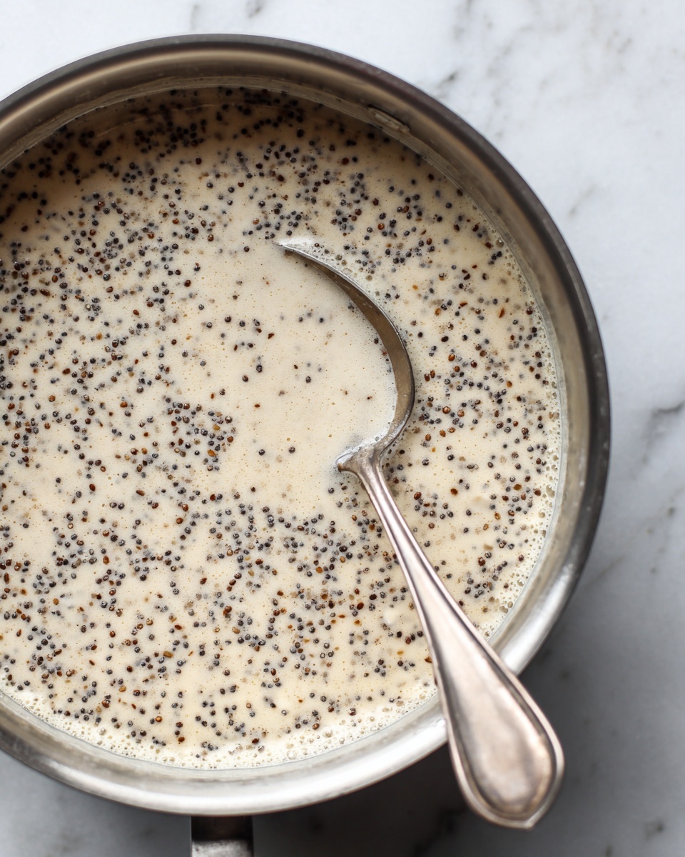 A close-up view of a large silver saucepan filled with a creamy mixture containing many small black seeds and brown bits evenly spread throughout the light beige liquid. A silver spoon is partially submerged in the mixture, its handle resting on the edge of the pot. The scene shows slight bubbles at the surface of the liquid, indicating gentle cooking. The pot sits on a stove with a white marbled surface visible below. photo taken with an iphone --ar 4:5 --v 7