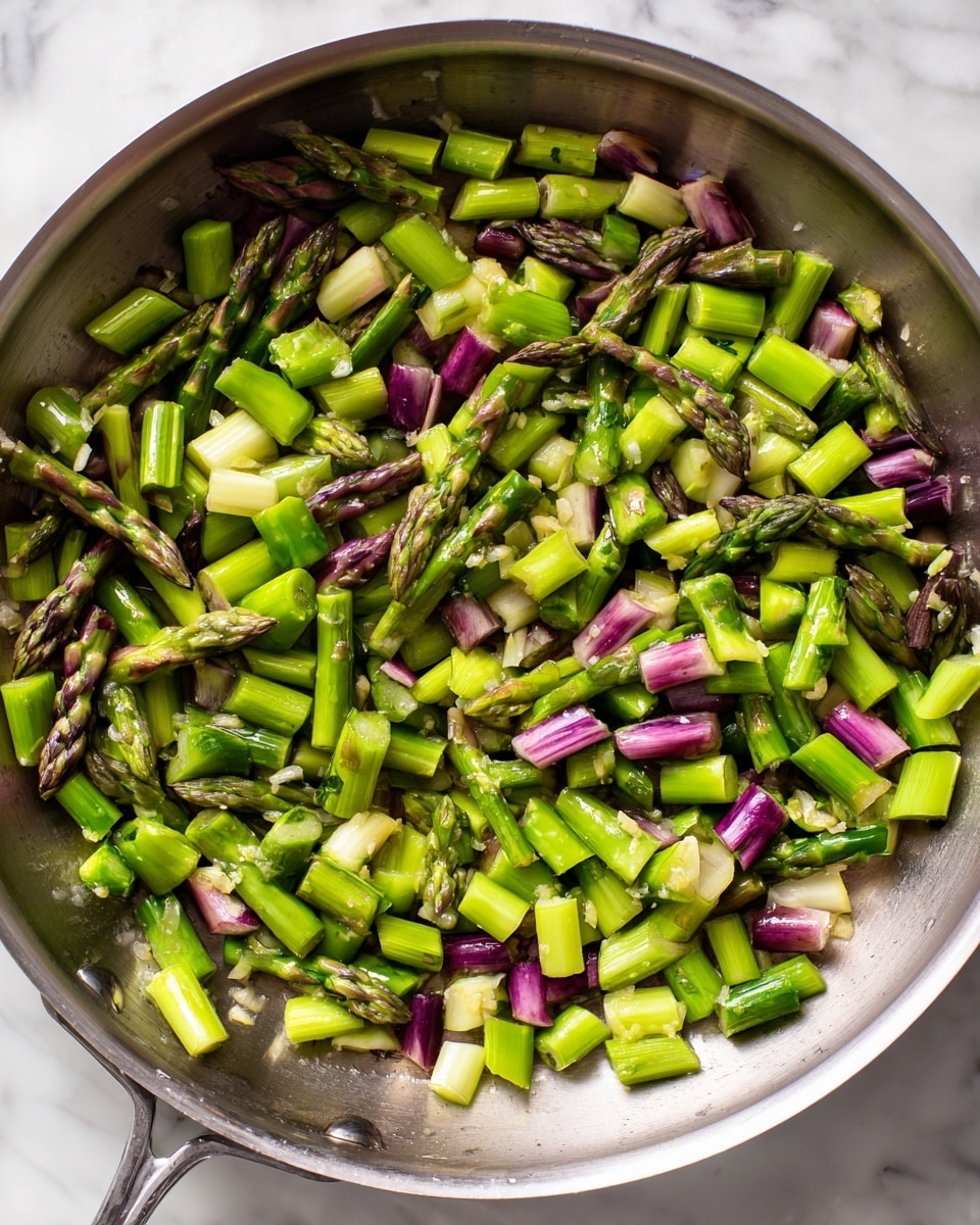 The image shows a close-up of chopped asparagus cooking in a silver pan. The asparagus pieces are cut into small chunks, with a mix of light green and purple tips scattered evenly across the pan. The pan's surface has a slight shine with melted butter and small bits of garlic visible under and around the asparagus. The colors are fresh and bright, with the asparagus green standing out against the shiny metal pan. The background is a white marbled texture. photo taken with an iphone --ar 4:5 --v 7