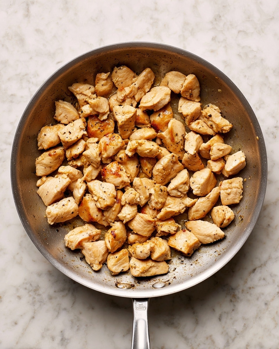A grey pan filled with small pieces of cooked chicken that are golden-brown on some sides, spread evenly inside the pan with light browning marks on the pan’s surface, all sitting on a white marbled textured background photo taken with an iphone --ar 4:5 --v 7