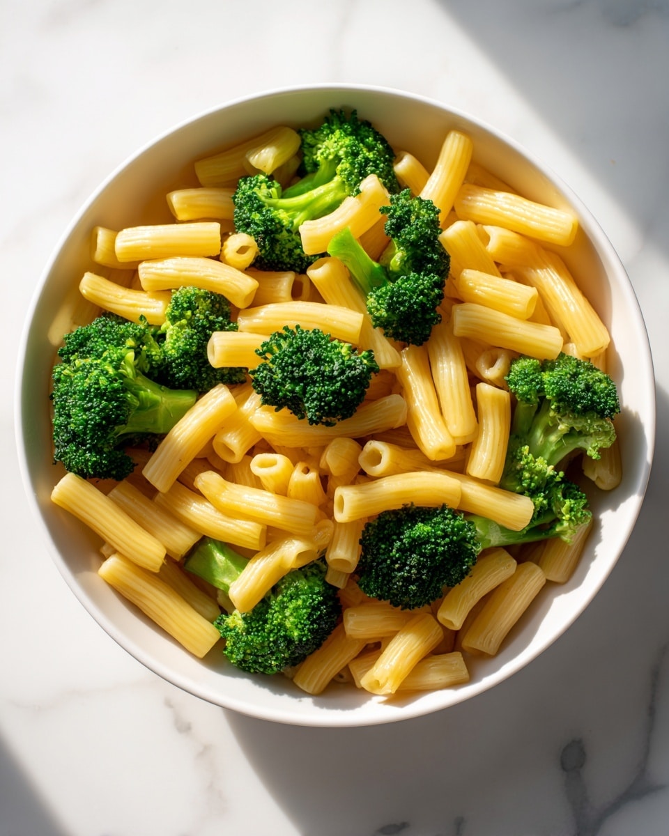 Inside a white bowl, there is a mix of short, yellow macaroni pasta and bright green broccoli florets. The pasta pieces are scattered all over, with some laying flat and others angled vertically. The broccoli pieces are chunky and spread evenly among the pasta, showing their textured, bumpy tops and solid stems. The bowl sits on a white marbled surface and the lighting creates a warm glow on the food, highlighting the fresh colors and slight shine on the pasta. photo taken with an iphone --ar 4:5 --v 7