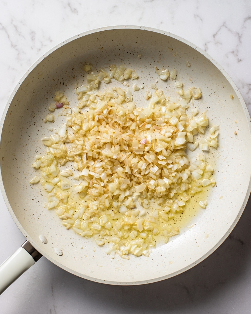 The image shows a close-up of a white pan with small chopped onion pieces being cooked in oil. The onions are pale yellow to light brown in color, with some pieces having a slight pink tint, and they are spread unevenly across the pan's surface. The oil around the onions shines, giving a glossy texture to the scene. The pan sits on a white marbled surface, giving a clean background. photo taken with an iphone --ar 4:5 --v 7