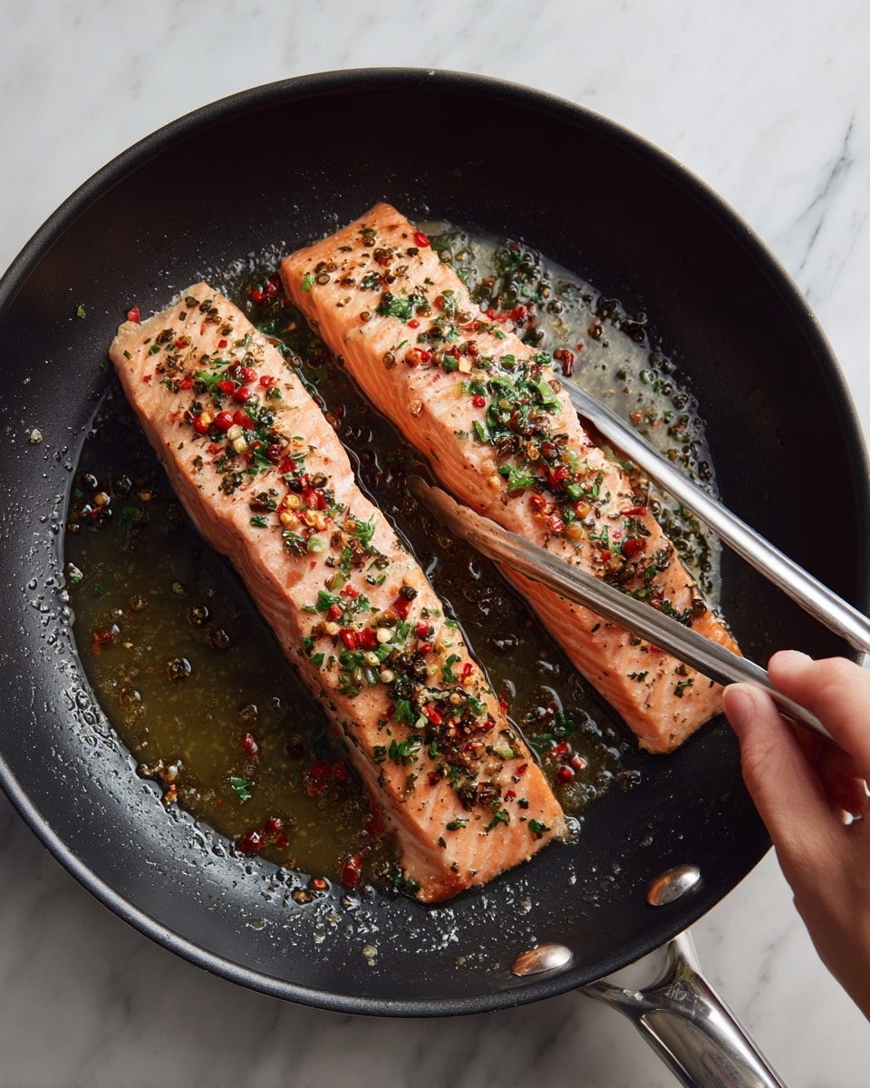 Two long, rectangular pieces of pink salmon are cooking in a black non-stick pan with some oil. The salmon has small bits of red chili flakes, black pepper, and green herbs sprinkled evenly on top. A pair of silver tongs held by a woman's hand is gently holding one piece of salmon. The pan sits on a white marbled surface. photo taken with an iphone --ar 4:5 --v 7