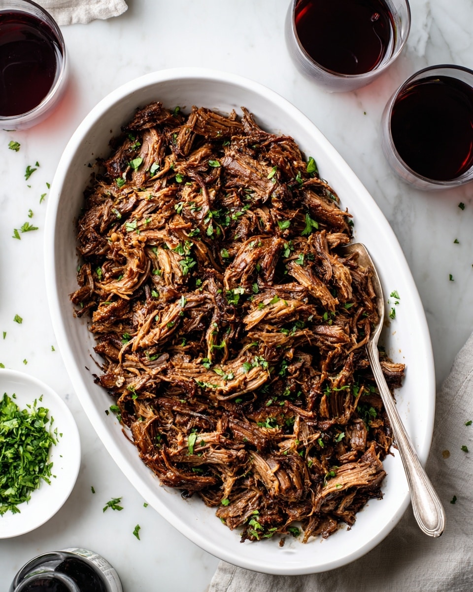 The image shows a white oval dish filled with shredded, cooked meat that is dark brown with some crispy edges and slight green herb sprinkles on top. A silver spoon is partially placed inside the dish on the right side, resting on the tender meat. The dish is on a white marbled surface, around which there are two glasses filled with a dark red liquid, a small white plate with chopped green herbs, and a dark bottle lying on its side near the bottom left corner. The overall setting is bright and clean, focusing on the rich texture and color contrast of the meat. photo taken with an iphone --ar 4:5 --v 7
