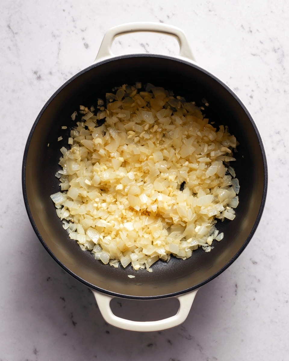 A top view of a black round pot with white handles placed on a white marbled surface. Inside the pot, there is a single layer of sautéed onion pieces that are translucent with a shiny, slightly oily texture. The onions are scattered unevenly, and there are tiny bits of minced garlic mixed in among the onion pieces. The pot has a matte black interior, contrasting with the light golden color of the cooked onions and garlic. Photo taken with an iphone --ar 4:5 --v 7