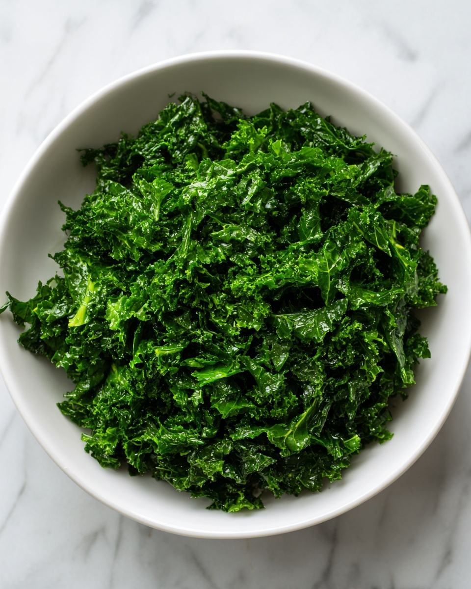 A white bowl filled with finely chopped dark green kale leaves, showing a dense, slightly wet texture as if lightly dressed or tossed. The kale pieces are layered evenly, covering the entire bowl with crisp edges visible. The bowl is set on a white marbled surface, enhancing the vibrant green color of the kale. Photo taken with an iphone --ar 4:5 --v 7