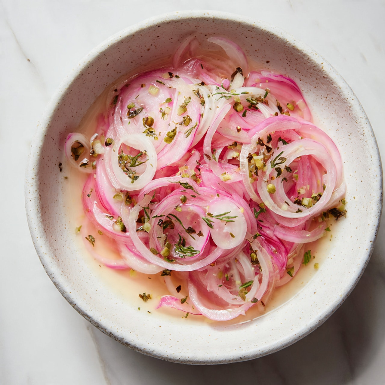 The image shows a dish of thinly sliced pink and white onions soaked in a light pink liquid, arranged loosely in a white speckled bowl with wide edges. The onions have a soft, translucent texture, layered unevenly with some pieces overlapping others. The liquid fills the bottom of the bowl, reflecting a slight shine. The bowl is placed on a white marbled surface with small green herbs and spice bits scattered around its edges. The overall color palette is soft with pinks, whites, and light reds, creating a fresh and simple look. photo taken with an iphone --ar 4:5 --v 7