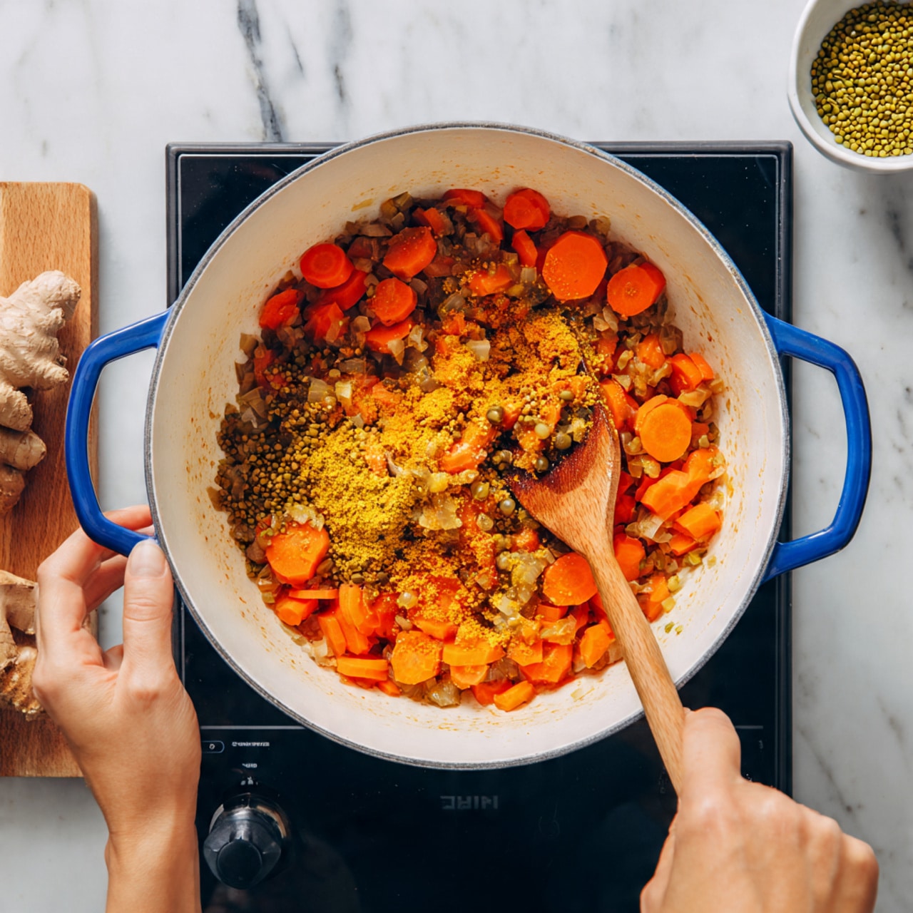 Inside a white pot with blue handles, there are cooked slices of orange carrots mixed with small chopped bits of brown onions and yellow spices. The food layers are scattered unevenly around the bottom of the pot, showing some spots with thicker patches of vegetables and seasoning. A wooden spoon is resting inside the pot with its handle held by a woman's hand on the right, stirring the mixture. Another woman's hand holds the pot handle on the left. The pot is placed on a black stove top, and the background surface is a white marbled texture. In the top right corner, there is a small white bowl filled with green mung beans and a piece of fresh ginger nearby. Photo taken with an iphone --ar 4:5 --v 7