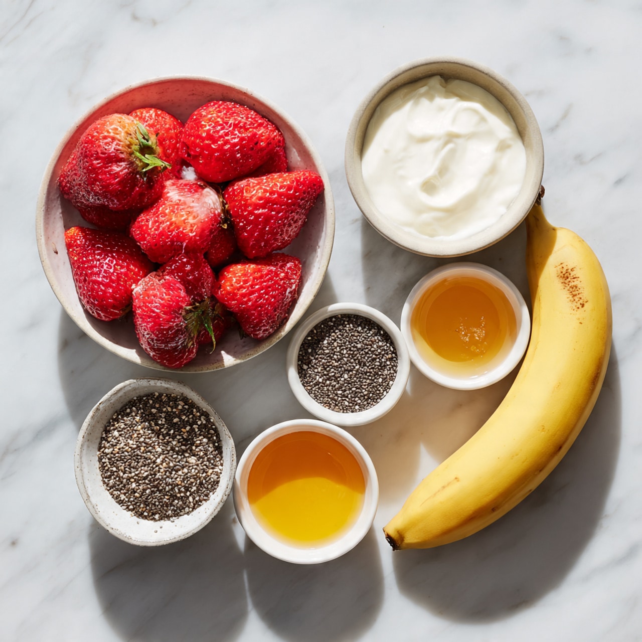 A clear glass blender jar is placed on a black base against a white marbled surface and blurred background. Inside the jar, the first layer at the bottom is white liquid, likely milk or yogurt. On top of that is a layer of vibrant red strawberry slices, some partially submerged in the milk. Above the strawberries, a sprinkling of tiny black chia seeds adds texture. The top layer consists of several light yellow banana slices that fill the jar up to the 1-liter mark. The blender jar has measurement markings on the side and a sturdy handle on the right. Photo taken with an iphone --ar 4:5 --v 7
