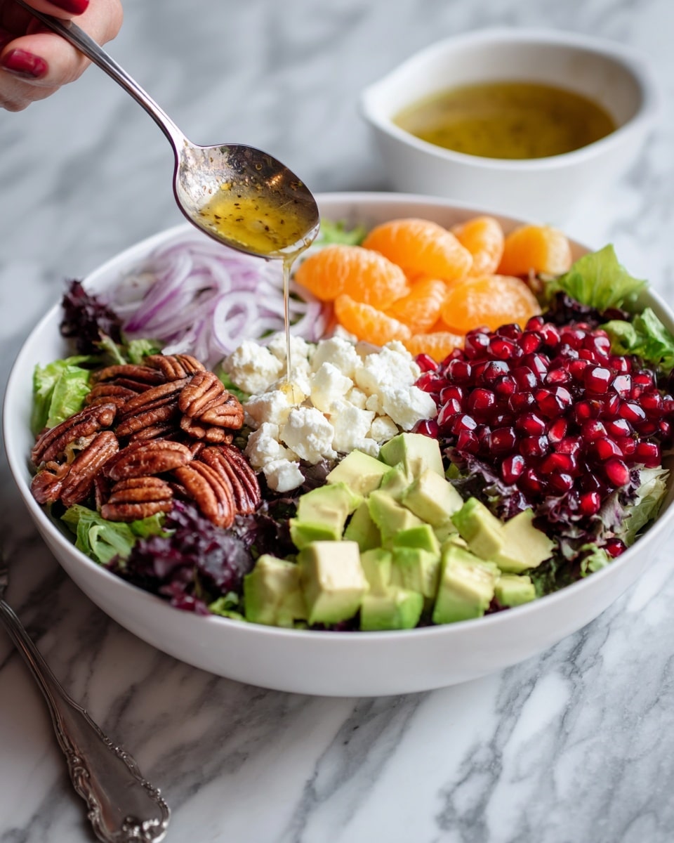 A white bowl filled with a layered salad starts with a base of mixed green and purple leafy lettuce covering the bottom. On top are piles of bright red pomegranate seeds, pale green cubed avocado on the right, glossy orange mandarin slices near the center, crumbled white cheese below them, thin white and purple sliced shallots on the left, and a cluster of candied pecans on the lower right. A woman's hand holds a spoon pouring a golden vinaigrette over the salad. Next to this bowl is a small white cup filled with the same vinaigrette, showing a shiny, smooth texture with herbs mixed in. The whole scene is set on a white marbled surface. Photo taken with an iphone --ar 4:5 --v 7
