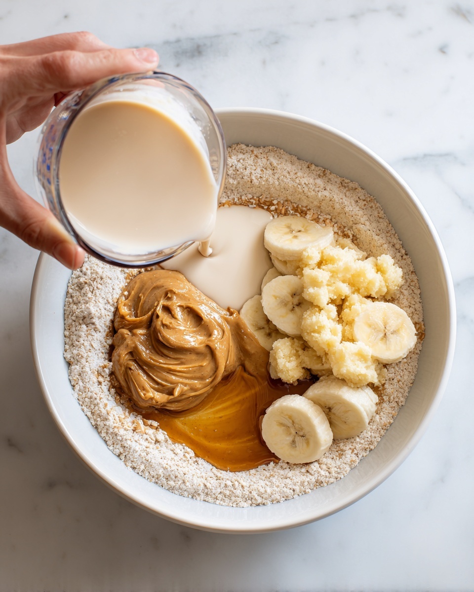 A white bowl contains a ring of light beige flour around the edges, with several ingredients placed separately inside the circle: a dollop of light brown peanut butter at the top center, thick golden syrup to the left, mashed pale yellow bananas next to the peanut butter, and a pool of creamy white liquid moving from the right towards the center. A woman's hand holds a clear measuring cup pouring more creamy white liquid into the bowl over a white marbled surface. Photo taken with an iphone --ar 4:5 --v 7