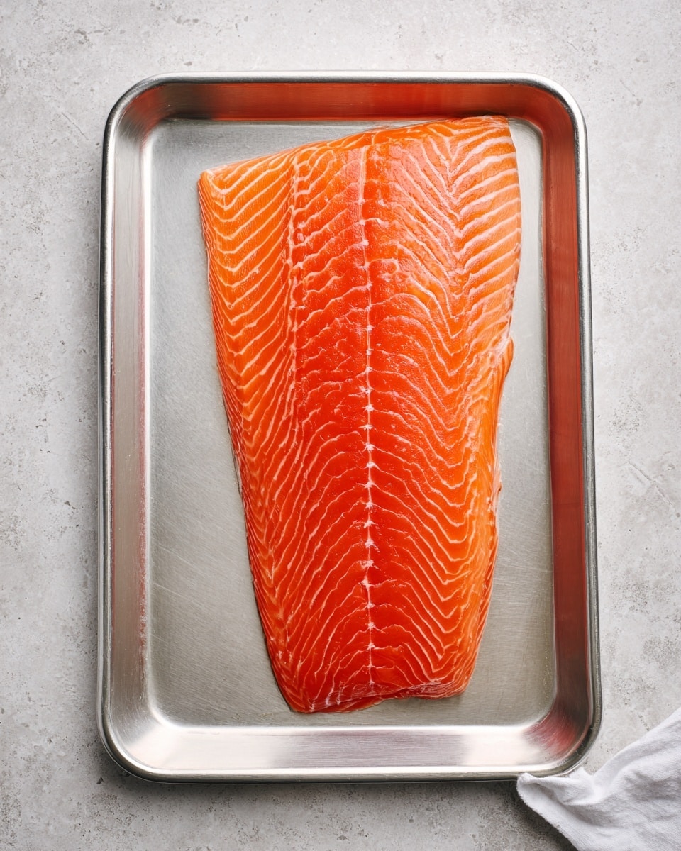 A large, raw salmon fillet with bright orange color and white fat lines is placed flat in a silver metal tray. The tray sits on a white marbled surface. A white cloth is slightly visible in the bottom right corner of the image. The salmon has smooth texture with clear, even layers. photo taken with an iphone --ar 4:5 --v 7