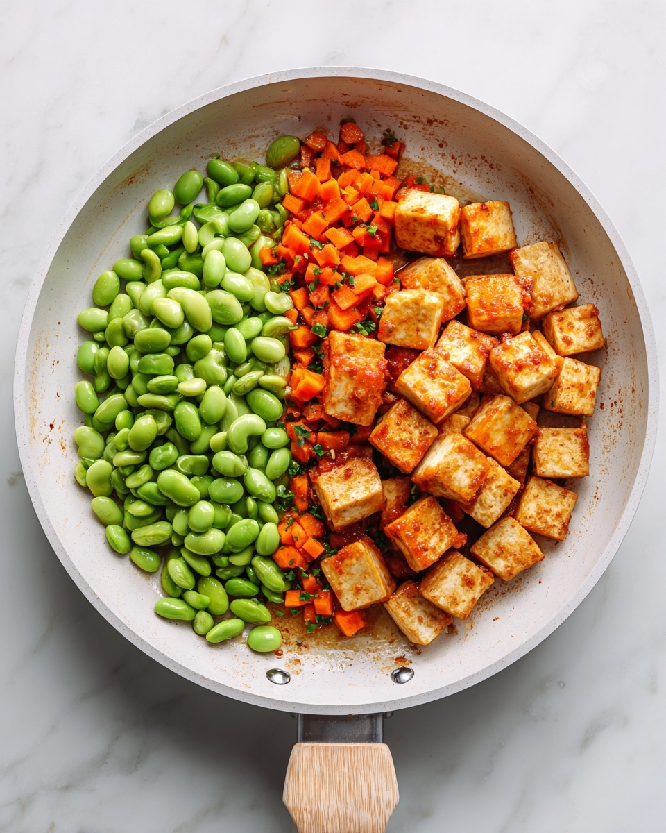 A white frying pan sits on a white marbled texture, filled with three main layers arranged side by side. On one side, there is a pile of bright green edamame beans with a smooth texture. Next to the beans, there is a layer of small orange carrot cubes mixed lightly with green herbs. The largest section contains golden-brown cubes of tofu, some topped with a reddish-orange sauce, giving it a slightly textured and sauced look. The pan handle is light wood-colored and points to the bottom left. The scene is lit naturally and clearly. photo taken with an iphone --ar 4:5 --v 7