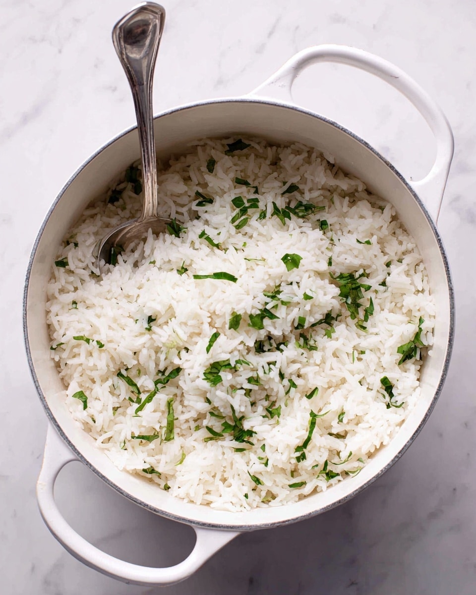 A white pot filled with cooked white rice mixed with small green herbs scattered evenly on top and throughout the rice. A silver spoon is placed inside the pot, partially buried in the rice on the left side. The pot is set on a white marbled surface, and the pot itself has a white handle. The texture of the rice looks soft and fluffy with the herbs providing a fresh contrast. photo taken with an iphone --ar 4:5 --v 7