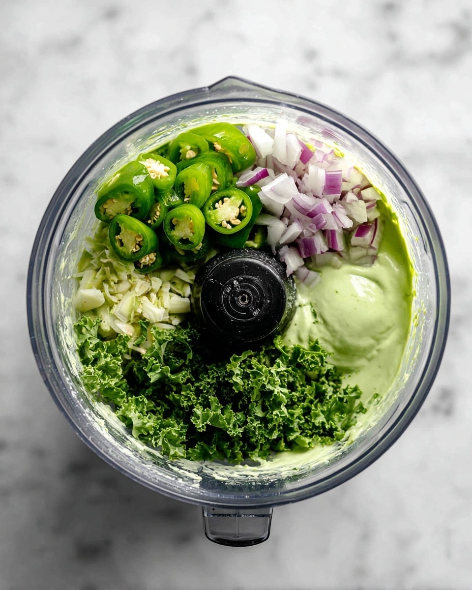Inside a clear blender jar on a white marbled surface, there is a layered mix of ingredients ready to be blended. The bottom layer is a smooth, light green creamy base that lines the sides of the jar. On top of this base are roughly chopped fresh green jalapeño pieces scattered next to bright purple and white chunks of onion. Around these, there are vibrant green leafy herbs and kale adding texture and depth. The sharp black blender blade sits prominently in the center, surrounded by the colorful fresh ingredients. photo taken with an iphone --ar 4:5 --v 7