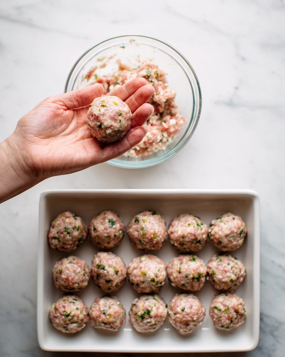 A woman's hand holds a small, round raw meatball made from a mix of ground meat and visible chopped herbs and onions, positioned above a clear glass bowl filled with the same mixture and a metal meat scoop resting inside. Next to it is a rectangular white baking dish arranged neatly with 15 similar raw meatballs, each showing the same mixed texture of meat, herbs, and onions, set on a white marbled surface. photo taken with an iphone --ar 4:5 --v 7