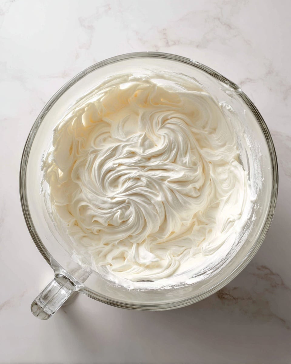A clear glass mixing bowl filled with smooth, thick white cream spread evenly in one layer, showing soft swirls and peaks on the surface. The bowl has a handle and sits on a white marbled texture surface. The cream looks rich and light, taking up about half the height of the bowl. Photo taken with an iphone --ar 4:5 --v 7