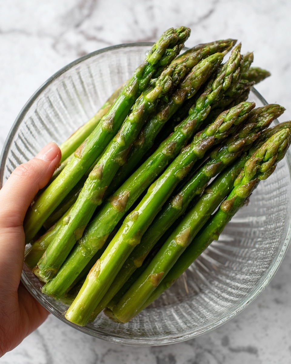 A close-up image shows a bunch of bright green asparagus spears that look slightly cooked or steamed. The asparagus spears have a smooth, glossy texture with some darker green tips. They are placed inside a clear glass bowl with subtle ridges. A woman's hand is gently holding several asparagus spears from the right side of the image. The background has a white marbled texture. Photo taken with an iphone --ar 4:5 --v 7