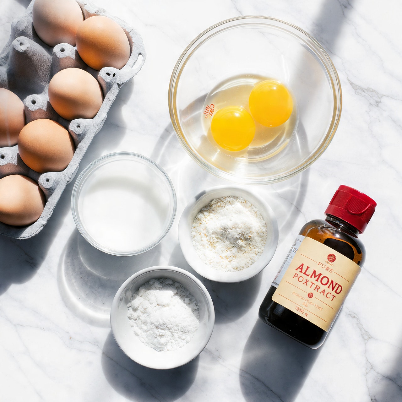 The image shows six brown eggs in a white egg carton on the left, with a clear glass bowl of yellow egg whites to the right of it. Below the eggs are three white bowls: one with a small amount of white liquid, one with a heap of white powder (likely sugar), and one with a smaller amount of white powder (likely baking powder). On the bottom right is a small dark bottle with a red cap and a label that reads