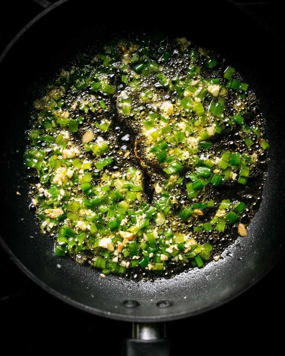 The image shows a close-up of a black frying pan on a stove with hot, bubbling oil inside. In the oil, there are small pieces of chopped green onions and white garlic, spread evenly across the pan's surface. The green onions are bright green with a fresh texture, while the garlic pieces are small and slightly golden. The bubbles from the oil create a shiny, lively texture all over the ingredients, and the pan’s black surface contrasts with the colors of the food. The background is dark, highlighting the frying pan. photo taken with an iphone --ar 4:5 --v 7