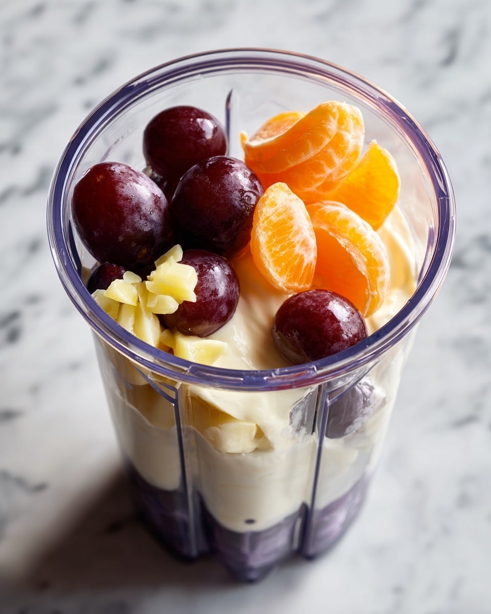 A clear blender container is filled with three visible layers: the bottom layer is made of whole dark purple grapes, the middle layer is creamy white yogurt that slightly drapes over the grapes, and the top layer consists of small light yellow chopped pieces and two peeled bright orange tangerine segments sitting on top. The blender sits on a white marbled surface. Photo taken with an iphone --ar 4:5 --v 7