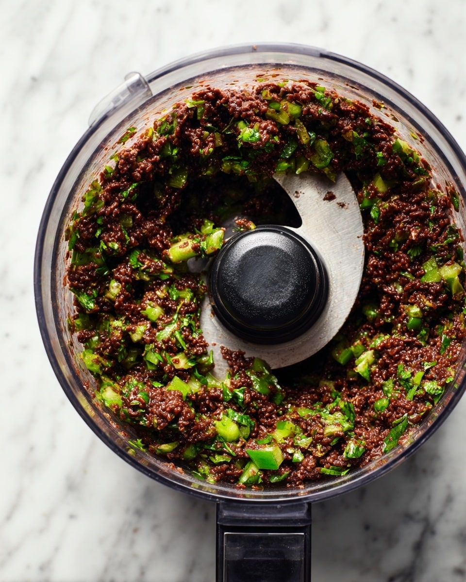 A top view of a clear food processor bowl filled with thick, chunky mixture of dark brown beans and bright green herbs, likely cilantro and possibly diced green peppers, mixed evenly throughout. The mixture clings to the sides of the bowl with some texture visible from the broken beans and leafy herbs. The black plastic blade sits in the center under the mixture. The bowl rests on a white marbled surface. photo taken with an iphone --ar 4:5 --v 7
