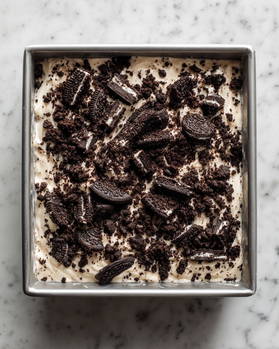 The image shows a square metal pan filled with a dessert that has two main layers. The bottom layer is a creamy white base mixed with small bits of crushed chocolate cookies, giving it a speckled look. On top, there is a generous scattering of larger chunks and crumbs of dark chocolate sandwich cookies, adding a rough texture to the smooth white surface. The metal pan is placed on a white marbled surface, highlighting the contrast between the dark cookie pieces and the light creamy base. Photo taken with an iphone --ar 4:5 --v 7