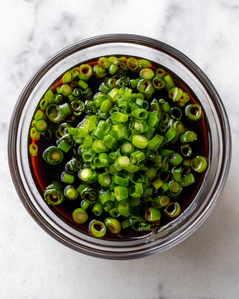 A clear glass bowl filled with dark soy sauce is topped with many small round chopped green onions floating on the surface, showing a mix of light and dark green colors. The bowl sits on a white marbled background that highlights the rich dark brown liquid and bright green onion pieces inside the bowl. photo taken with an iphone --ar 4:5 --v 7