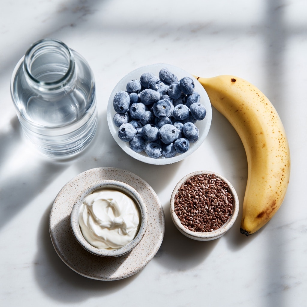 A clear blender jar with visible measurement marks holds three layers: the bottom layer is dark blue blueberries, the middle layer is a peeled banana cut in half showing its pale creamy texture, and the top layer is a dollop of white cream sprinkled with small dark brown chocolate shavings. The blender base is black with silver accents and a round central dial with buttons around it, standing on a white marbled surface with a plain white wall background. photo taken with an iphone --ar 4:5 --v 7