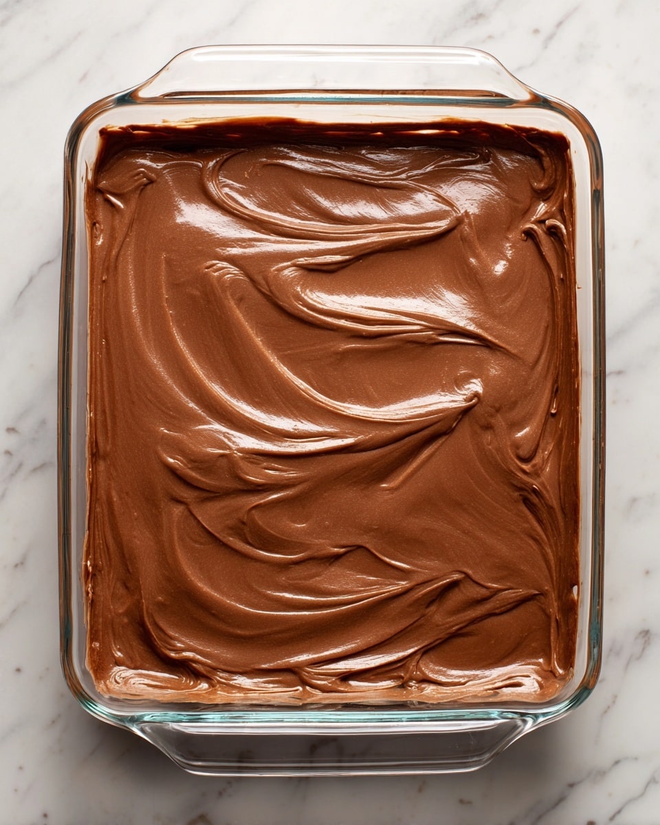 A clear glass baking dish sits on a white marbled surface, filled with a thick, smooth layer of shiny light brown chocolate batter that has been spread evenly, showing gentle swirls and a slightly glossy texture on top. The batter covers the entire flat base of the dish, with the edges touching the sides but not overflowing. Photo taken with an iphone --ar 4:5 --v 7