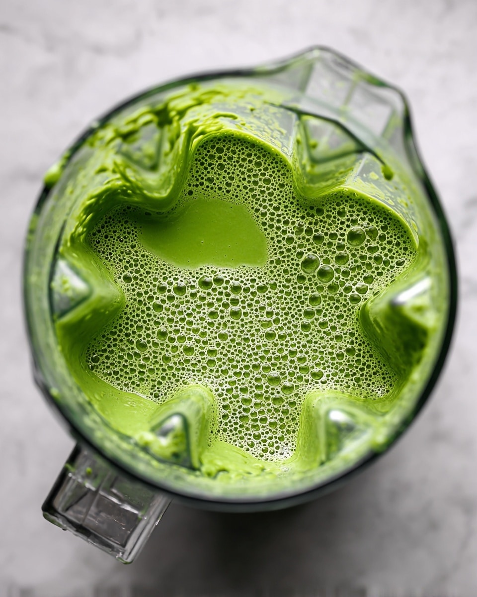 A clear blender jar filled with a bright green smoothie showing many small bubbles and foam on the surface, giving it a light, airy texture. The blender is on a white marbled surface, and the edges of the blender jar are slightly messy with some smoothie splashes. The green color is vivid and uniform, with no visible chunks. Photo taken with an iphone --ar 4:5 --v 7