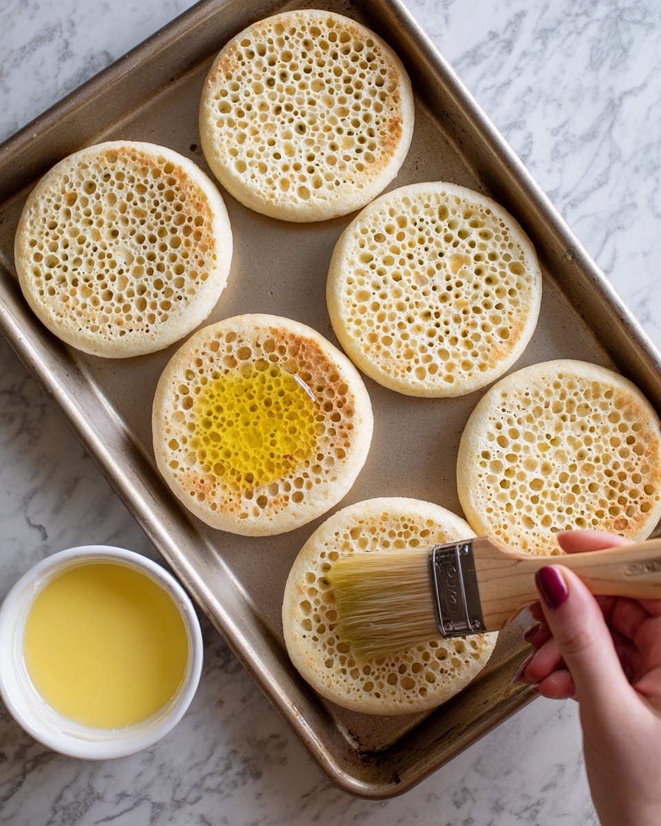 Six pieces of crumpets with a light golden color and many small holes are spread out on a metal baking tray. The crumpets are soft with a spongy texture. A woman's hand is holding a wooden brush, lightly spreading yellow melted butter on one crumpet at the bottom left. Below the tray, there is a white bowl filled with more melted butter. The whole setup is on a white marbled surface. photo taken with an iphone --ar 4:5 --v 7