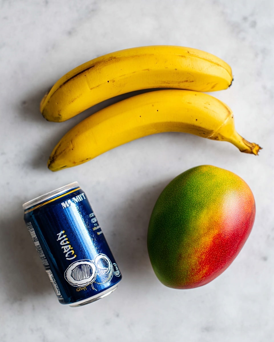 The image shows three ingredients placed on a white marbled surface: two peeled bananas positioned close together at the top, a whole mango on the bottom right with green, yellow, and red colors blending on its skin, and a dark blue can of unsweetened coconut milk on the bottom left, tilted with the label showing a coconut illustration and white text. The arrangement is simple and clean, with the fruits and can spaced evenly for a clear view photo taken with an iphone --ar 4:5 --v 7